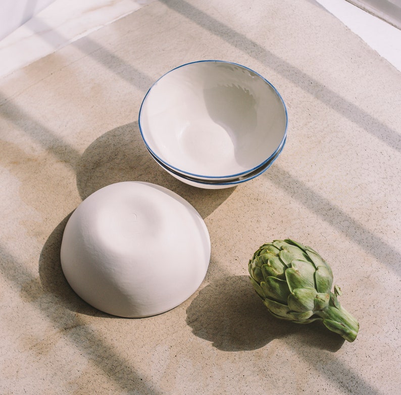 May include: Two white ceramic bowls, one with a blue rim, and a green artichoke on a light brown surface.