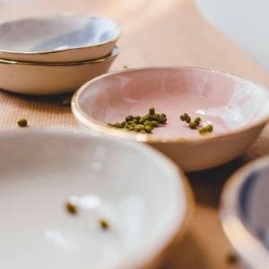 Może przedstawiać: Cztery małe ceramiczne miski z pozłacanymi brzegami. Miski są w r&oacute;żnych kolorach, w tym r&oacute;żowym, białym i niebieskim. Jedna miska zawiera zielone fasolki mung.