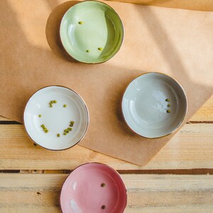 May include: Three small bowls with gold rims, each filled with a few green mung beans. The bowls are green, gray, and pink. The bowls are on a brown paper surface over a wooden surface.