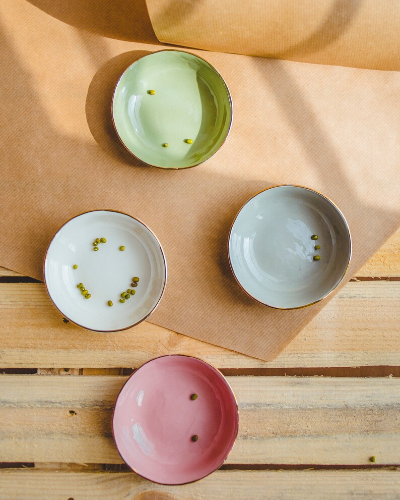 May include: Three small bowls with gold rims, each filled with a few green mung beans. The bowls are green, gray, and pink. The bowls are on a brown paper surface over a wooden surface.