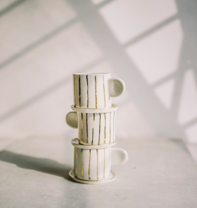 May include: A stack of three white ceramic coffee mugs with gold stripes. The mugs are stacked on top of each other and have a simple, elegant design.