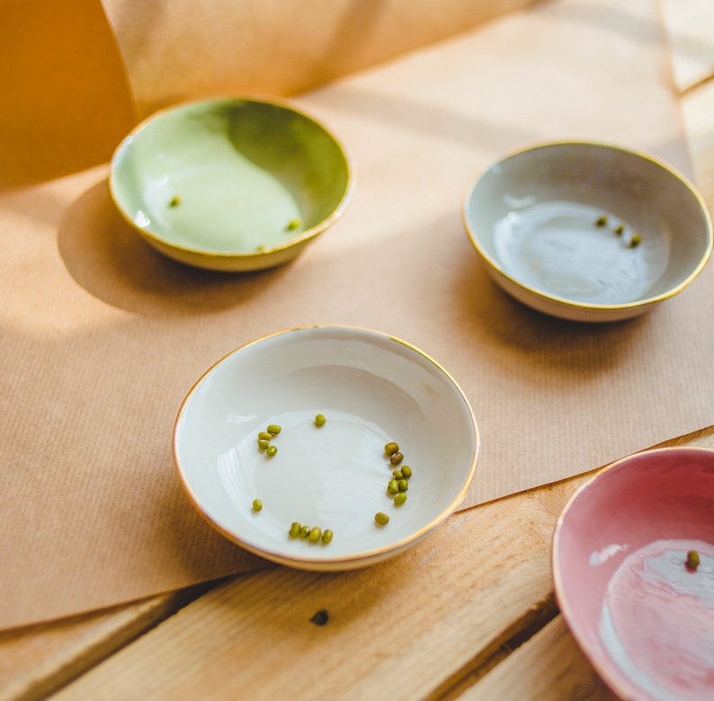 May include: Four small ceramic bowls with gold rims, three of which are filled with green mung beans. The bowls are on a brown paper surface and a wooden surface.