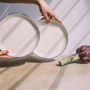 May include: Two white ceramic plates with a blue rim, one stacked on top of the other, and an artichoke on a light brown surface.