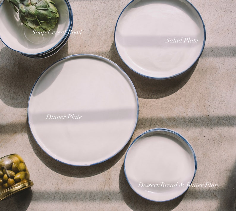 May include: A set of four white ceramic plates with blue trim. The plates are arranged in a circle, with the largest plate in the center and the smallest plate on the top. The plates are labeled "Dinner Plate", "Salad Plate", "Soup/Cereal Bowl", and "Dessert/Bread & Butter Plate".