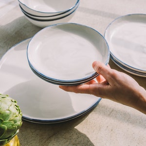 May include: A set of white ceramic plates with a blue rim. The plates are stacked on top of each other. The plates are on a light brown surface.