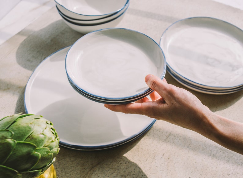 May include: A set of white ceramic plates with a blue rim. The plates are stacked on top of each other. The plates are on a light brown surface.