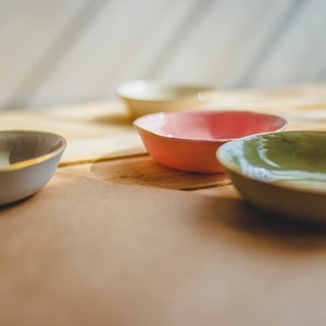 May include: Four small ceramic bowls in different colors: light blue, light green, pink, and cream. The bowls are arranged on a wooden surface.