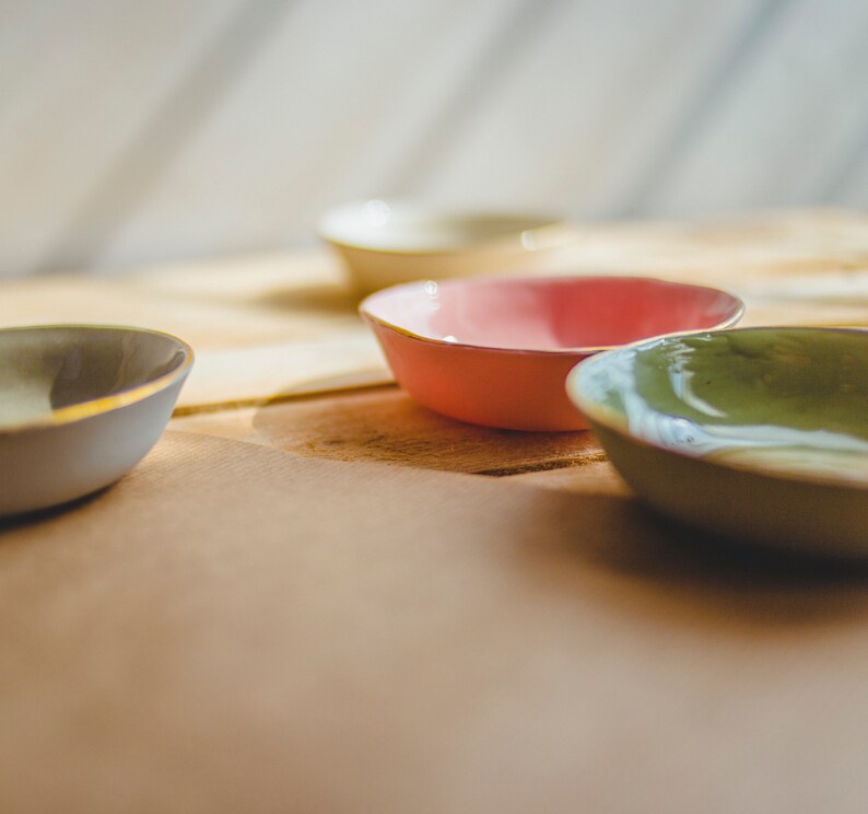 May include: Four small ceramic bowls in different colors: light blue, light green, pink, and cream. The bowls are arranged on a wooden surface.