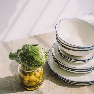 May include: A green artichoke sits on top of a glass jar filled with green olives. The jar is on a light gray surface. Behind the jar are a stack of white plates with blue trim.