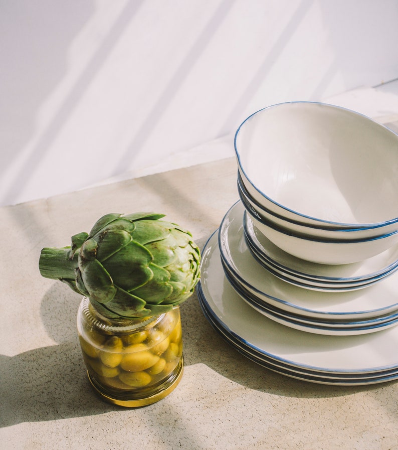 May include: A green artichoke sits on top of a glass jar filled with green olives. The jar is on a light gray surface. Behind the jar are a stack of white plates with blue trim.