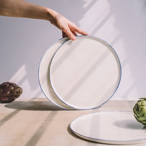 May include: Two white ceramic plates with a blue rim, one stacked on top of the other. The plates are on a light brown surface with a green artichoke in the foreground.