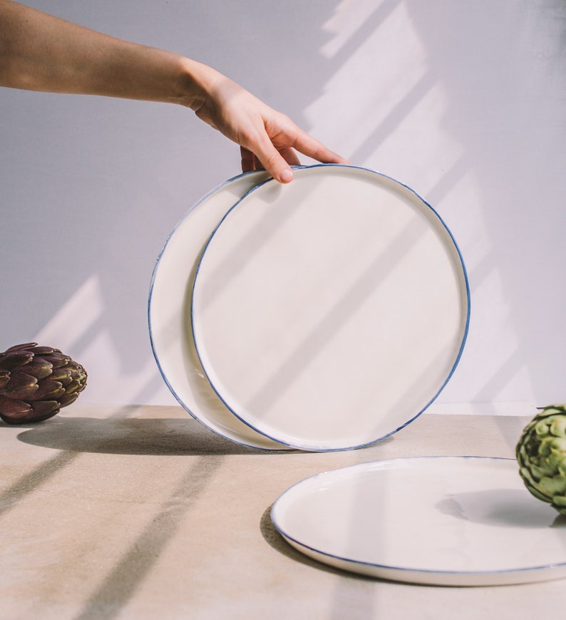 May include: Two white ceramic plates with a blue rim, one stacked on top of the other. The plates are on a light brown surface with a green artichoke in the foreground.