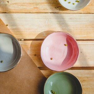 May include: Four small bowls with gold rims, each containing a few green mung beans. The bowls are on a wooden surface, with one bowl on a brown paper surface.