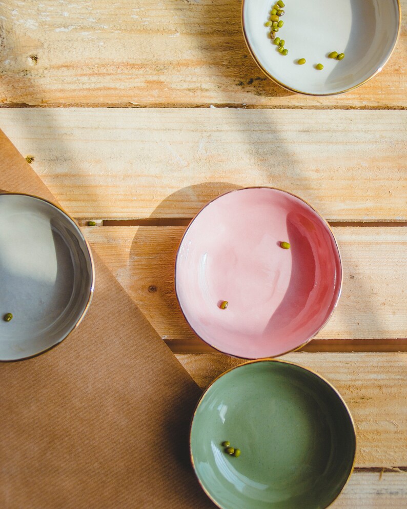 May include: Four small bowls with gold rims, each containing a few green mung beans. The bowls are on a wooden surface, with one bowl on a brown paper surface.