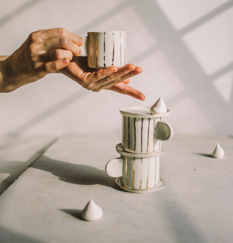 May include: A set of three white ceramic cups with gold stripes stacked on top of each other. The cups are on a gray surface with a white meringue on top of the top cup.
