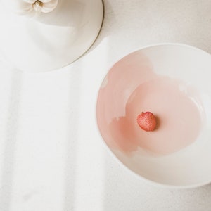 May include: A white bowl with a pink interior and a single lychee fruit in the center.