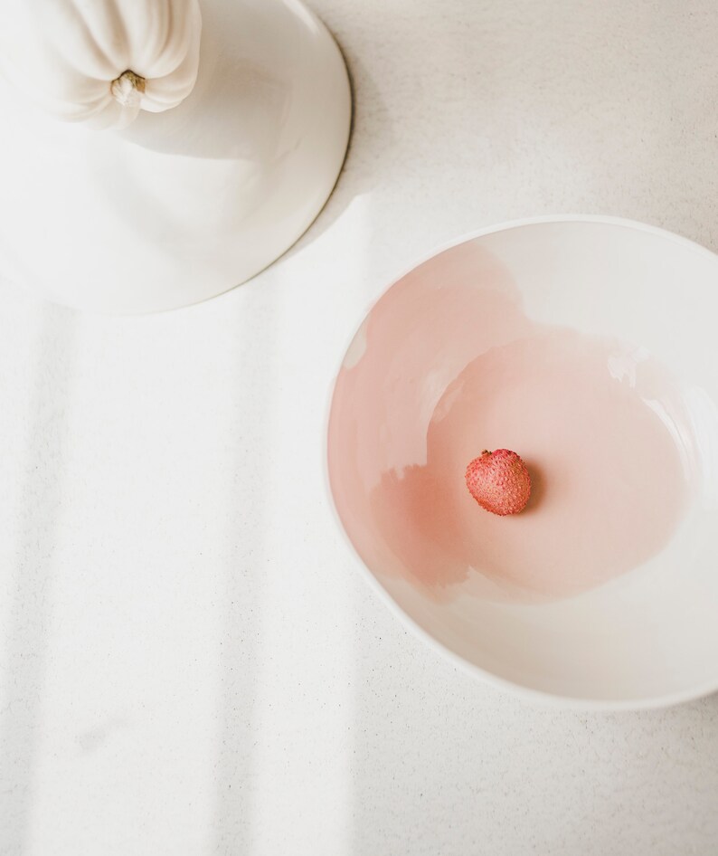 May include: A white bowl with a pink interior and a single lychee fruit in the center.