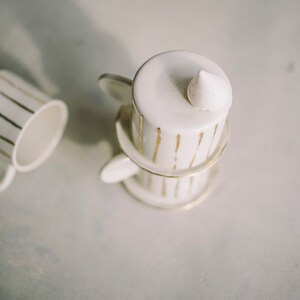 May include: A white teacup and saucer set with gold stripes. The teacup has a small white ceramic piece on top.