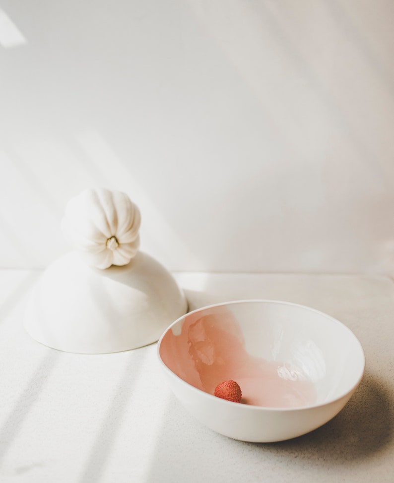 May include: A white ceramic bowl with a pink glaze on the inside. A single red lychee sits in the bowl.