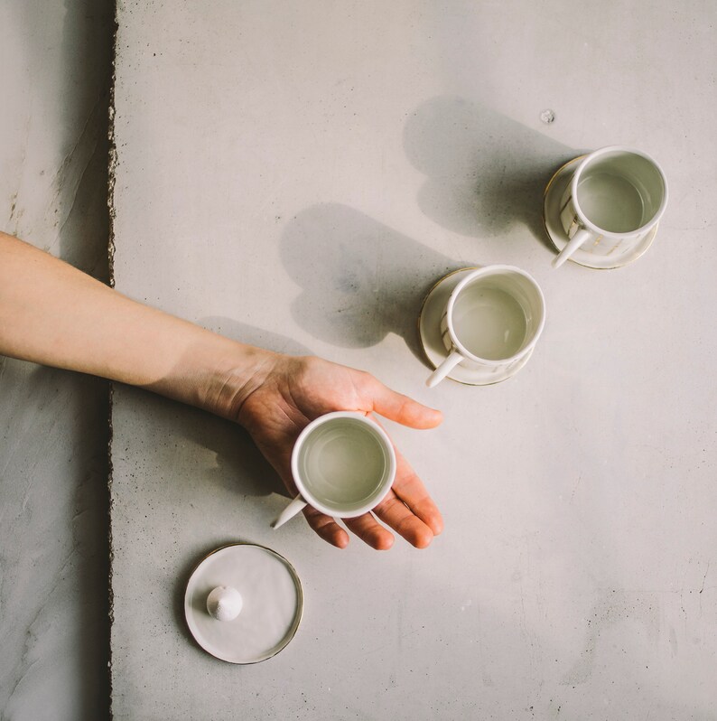 May include: Four white ceramic teacups with gold accents. Three teacups are on a white surface, and one is being held in a hand. The teacups have a simple design and are perfect for a relaxing afternoon tea.