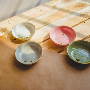 May include: Four small bowls with a light brown, gray, pink, and green glaze. The bowls are arranged on a wooden surface with a brown paper sheet underneath.