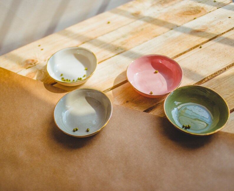 May include: Four small bowls with a light brown, gray, pink, and green glaze. The bowls are arranged on a wooden surface with a brown paper sheet underneath.