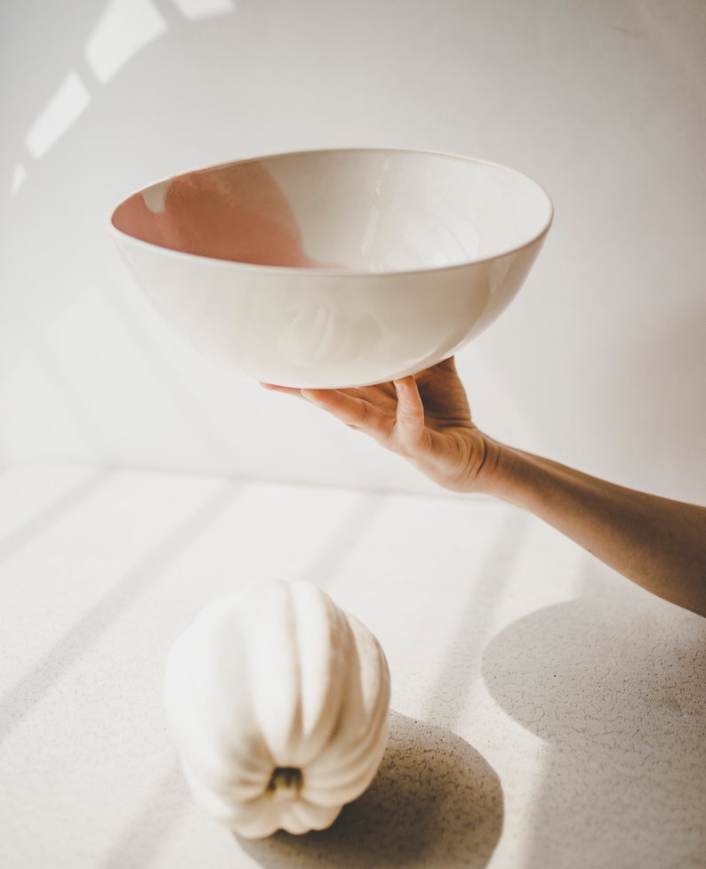 May include: A white ceramic bowl with a pink rim is held in a hand against a white background. A white pumpkin sits on the surface below the bowl.