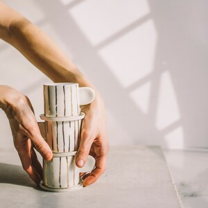 May include: A stack of three white ceramic mugs with gold stripes. The mugs are stacked on top of each other and are being held by a person's hands.