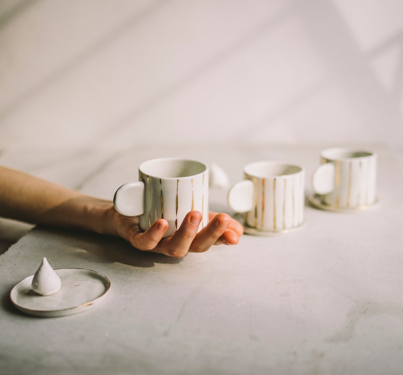 May include: Three white ceramic mugs with gold stripes. The mugs have a unique, rounded handle. One mug is being held in a hand.