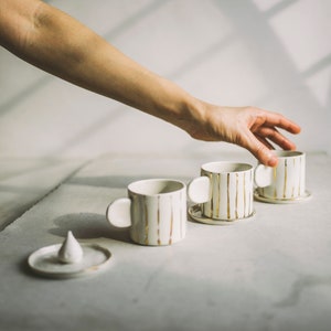 May include: Three white ceramic mugs with gold stripes and rounded handles. The mugs are on white saucers and are arranged in a row on a white surface.