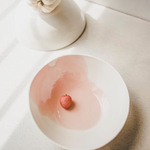 May include: A white bowl with a pink interior and a single red lychee fruit in the center.