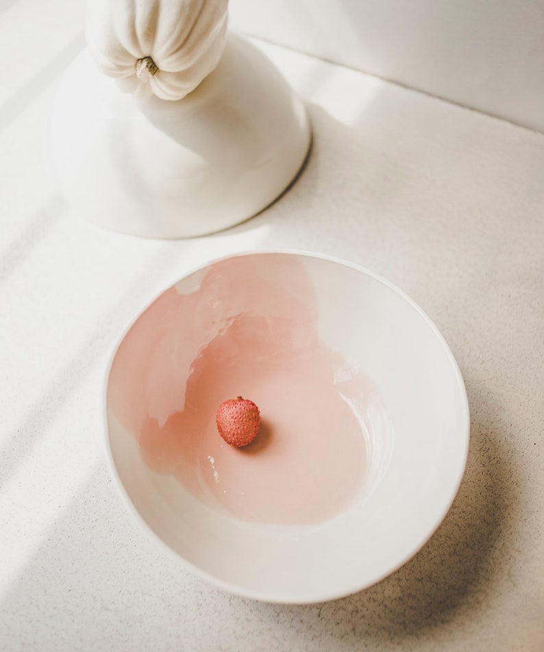 May include: A white bowl with a pink interior and a single red lychee fruit in the center.