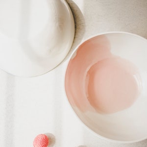 May include: Two white ceramic bowls, one with a pink interior, on a white surface with two red lychee fruits.