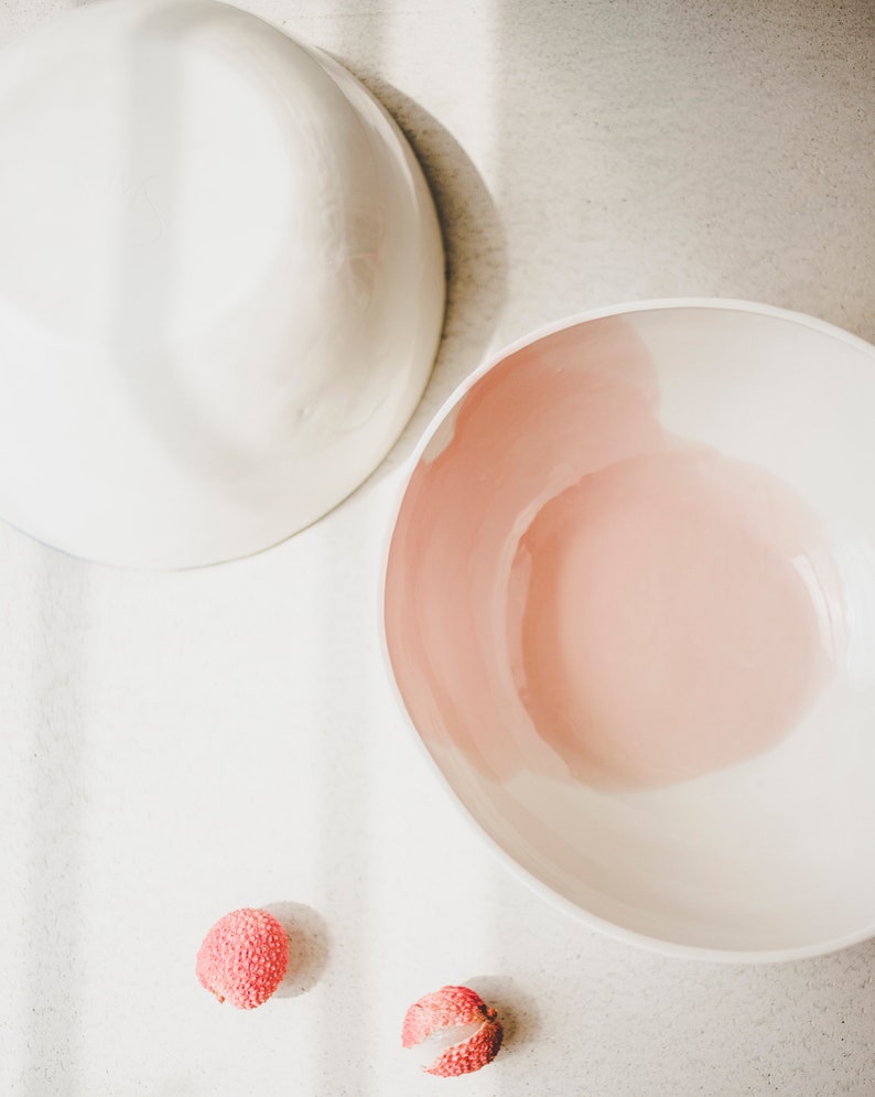 May include: Two white ceramic bowls, one with a pink interior, on a white surface with two red lychee fruits.