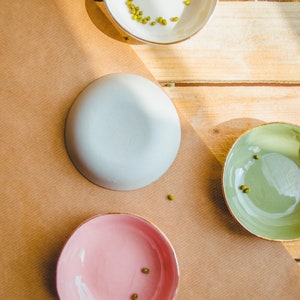 May include: Three small bowls with gold rims. The bowls are white, light blue, and pink. Each bowl has a few green mung beans inside.