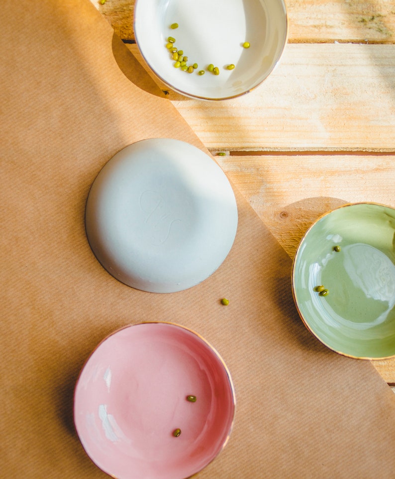 May include: Three small bowls with gold rims. The bowls are white, light blue, and pink. Each bowl has a few green mung beans inside.
