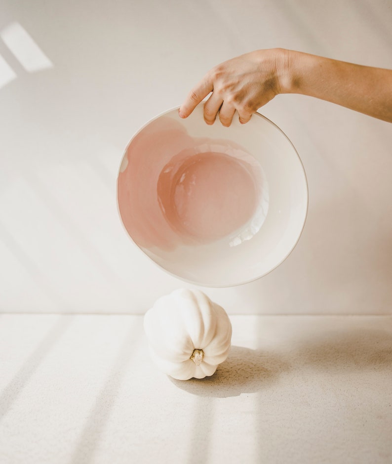 May include: A white ceramic bowl with a pink interior is being held upside down over a white gourd. The bowl and gourd are on a white surface.
