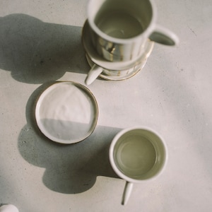 May include: A set of three white ceramic mugs with gold accents stacked on top of each other. The mugs are on a gray surface with a white saucer next to them.