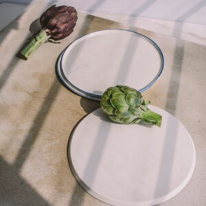May include: Two white ceramic plates with blue trim, one stacked on top of the other. A green artichoke sits on the top plate, and a purple artichoke sits on a light brown surface.