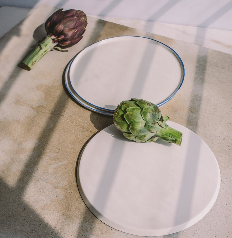 May include: Two white ceramic plates with blue trim, one stacked on top of the other. A green artichoke sits on the top plate, and a purple artichoke sits on a light brown surface.
