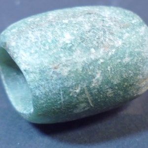 Könnte beinhalten: Eine verwitterte, zylindrische Perle mit einem Loch in der Mitte. Die Perle hat eine gedämpfte teal Farbe mit einer rauen, strukturierten Oberfläche. Die Form ist abgerundet, mit einer leicht unregelmäßigen Form, was darauf hindeutet, dass sie handgefertigt oder natürlich ist.