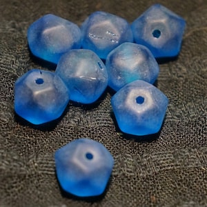 May include: A collection of eight faceted, light blue glass beads. Each bead has a pentagonal shape with a hole drilled through the center. The beads have a frosted appearance and are scattered on a dark, textured surface.