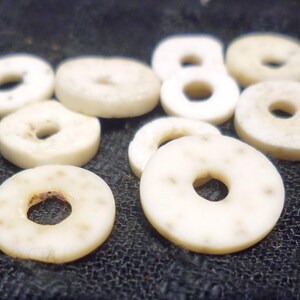 May include: A collection of white, round, shell beads with a hole in the center. The beads are arranged in a pile on a dark fabric surface.