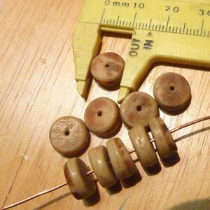 Puede incluir: Primer plano de un conjunto de ocho cuentas de madera marrón y blanco con un agujero en el centro de cada cuenta. Las cuentas están enhebradas en un alambre de cobre. Una pinza de plástico amarilla está midiendo una de las cuentas.