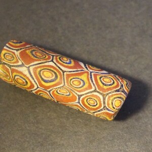 Op de afbeelding: Een cilindrische glazen kralen met een geometrisch patroon van rode, gele, blauwe en witte cirkels en zeshoeken.