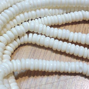 以下が含まれることがあります： 白い貝殻のビーズの糸。各ビーズは小さく、丸く、わずかに平らな円盤形です。ビーズは糸でつなぎ合わせて、長いネックレスになっています。