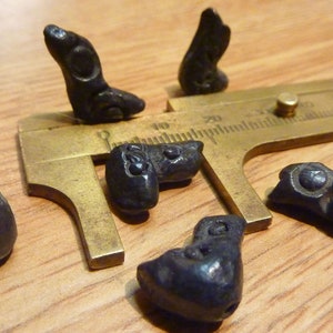 May include: Six black ceramic beads with intricate designs, some resembling animal figures, are arranged on a wooden surface with a brass measuring tool.
