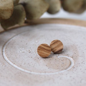 May include: Two small, round wooden stud earrings with a striped grain pattern. They are presented on a speckled, off-white ceramic plate with a spiral design. Dried eucalyptus leaves are in the background.