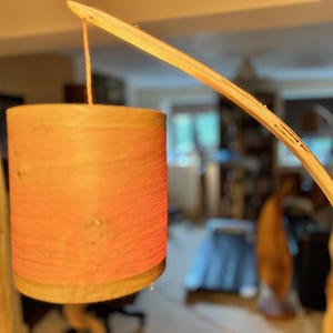 May include: A wooden lamp with a curved arm and a cylindrical shade made of woven material. The shade is a light orange color and has a natural wood base.