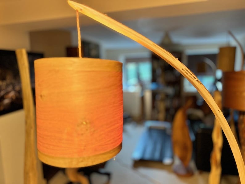 May include: A wooden lamp with a curved arm and a cylindrical shade made of woven material. The shade is a light orange color and has a natural wood base.